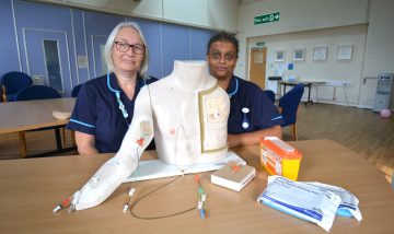 Rebecca McGlone and Jaimini Amin in their nursing uniforms, standing behind a desk with a mannequin torso used to practice procedures.