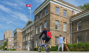 Image of Leicester University buildings on a sunny day, with students walking and on a bicycle