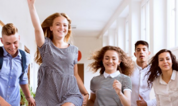 five teenagers in school uniform run down a corridor. One is jumping with her hand in the air.