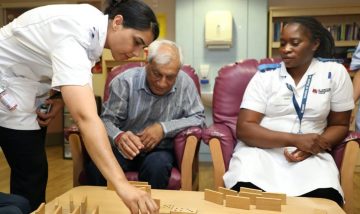 An older man sat between two nurses, he is leaning forward to place a domino on a table.