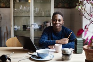 A man in a blue jumper sat at a table with his laptop and holding a pencil. He is looking at the camera and smiling with a china cup next to him.