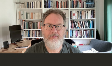 White middle aged man, brown hair, white beard, in front of book shelves