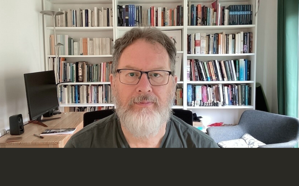 White middle aged man, brown hair, white beard, in front of book shelves