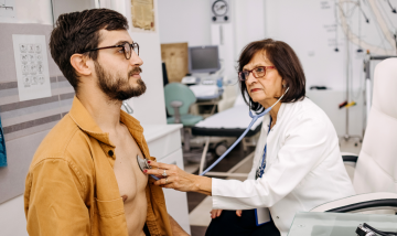 white middle aged female doctor examines man with beard in 30s using a stethoscope