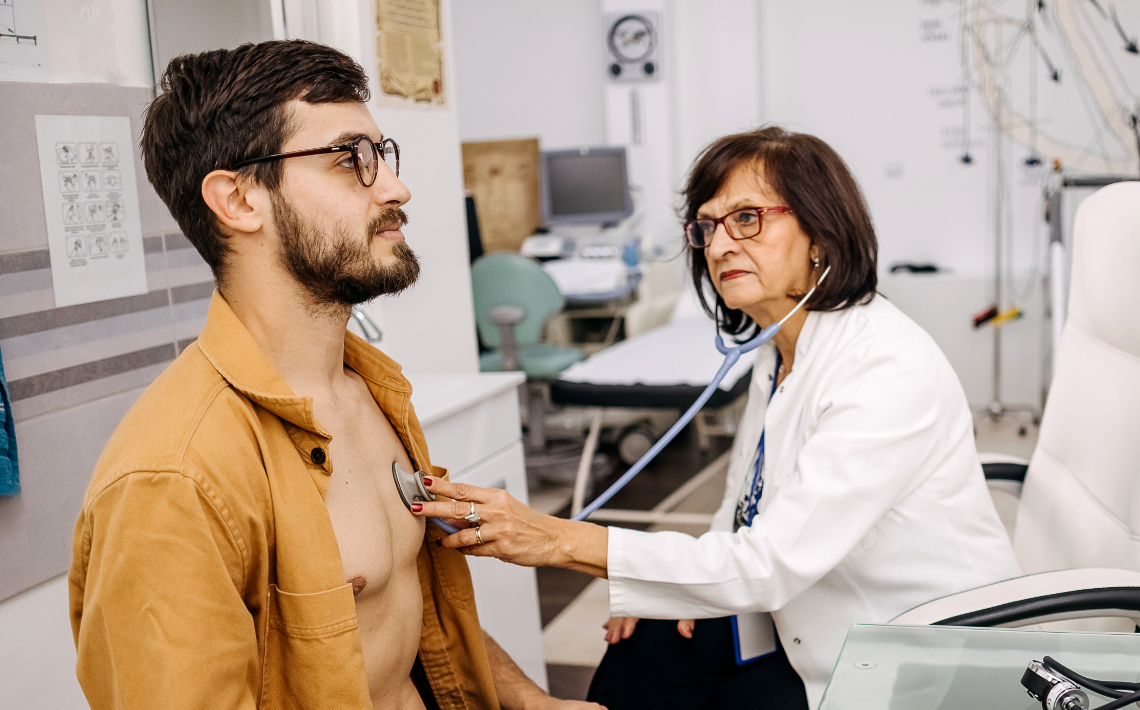 white middle aged female doctor examines man with beard in 30s using a stethoscope
