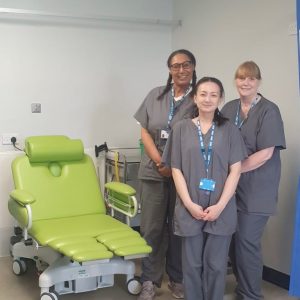 Three clinical staff stood smiling at the camera, to their left is a large green chair for sitting and lying in while receiving treatment.