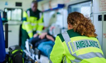 Ambulance interior focusing on the back of a paramedic comforting patient being transported to hospital.