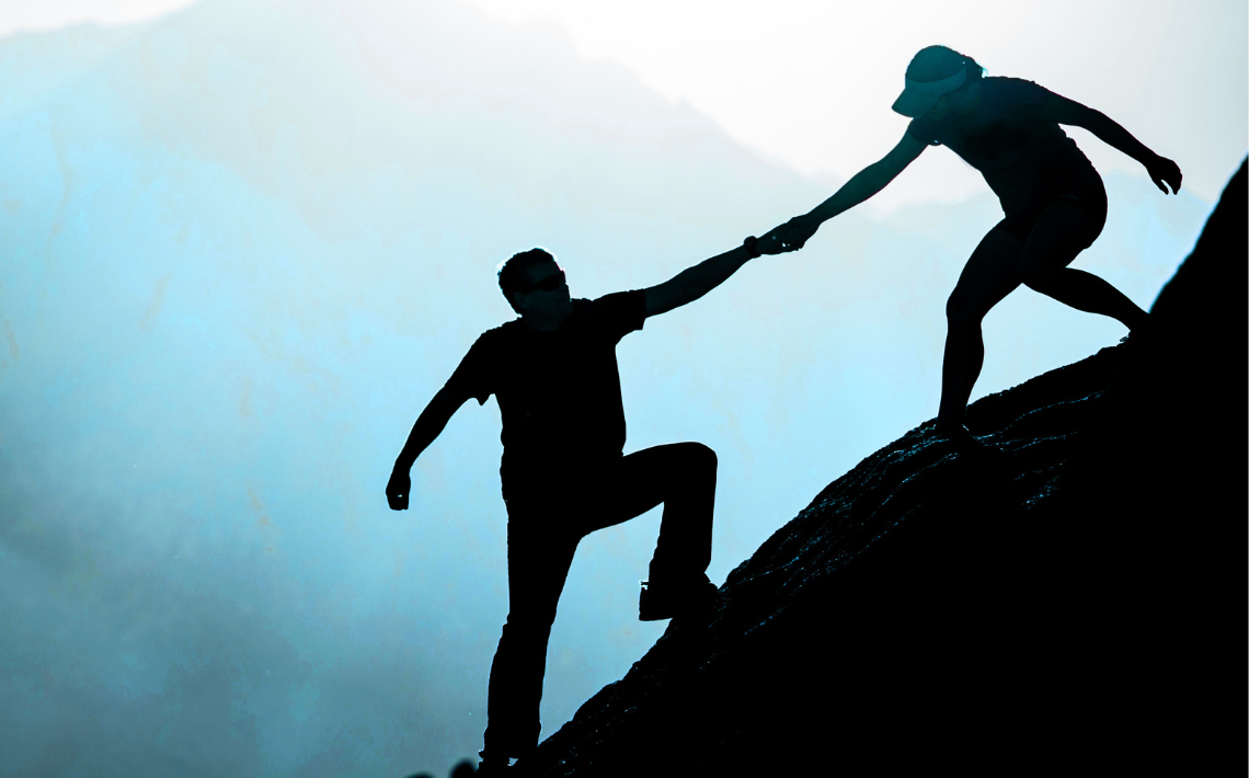 silhouettes of two people on mountainside - one person helping the other by pulling them by the hand