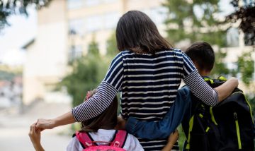 mother with her arms round a child on each side, walking away. each child is wearing a rucksack