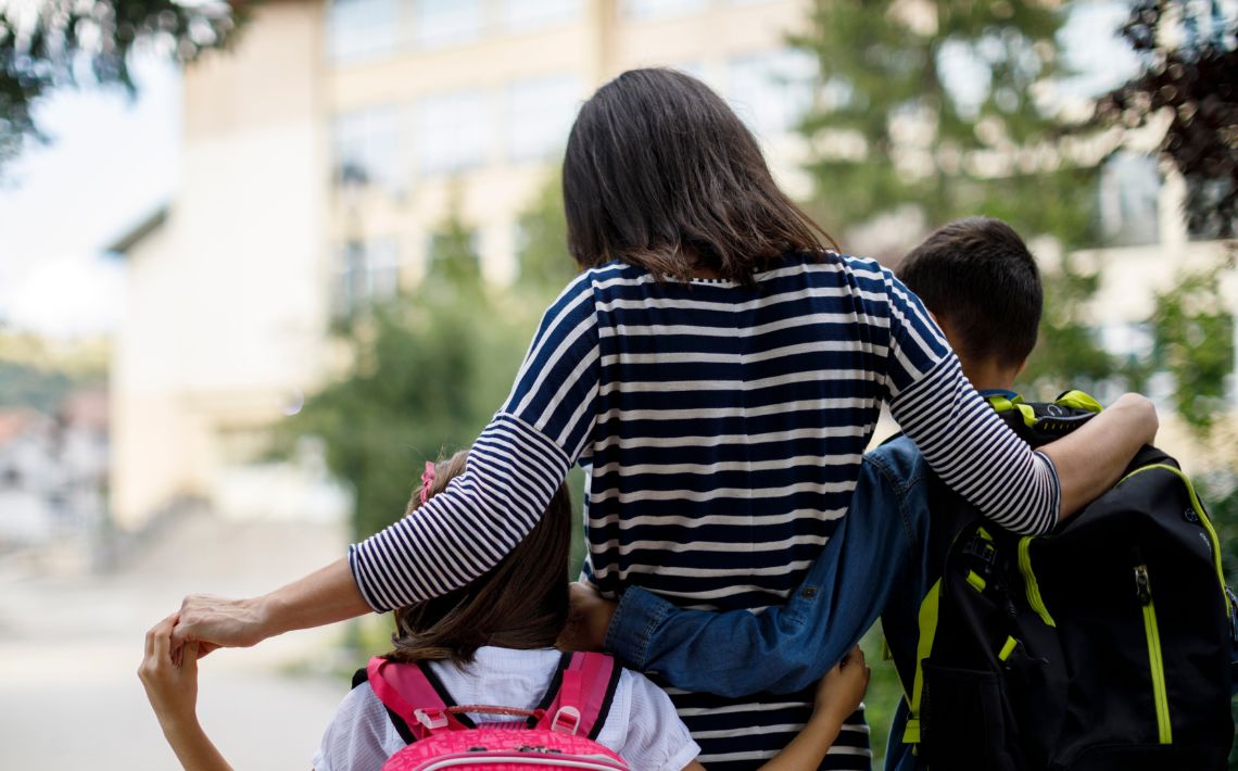 mother with her arms round a child on each side, walking away. each child is wearing a rucksack