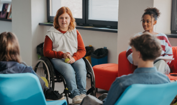 young people sitting in a circle - one white girl in a wheel chair, one black girl with hair up, white person unknown gender with back to view