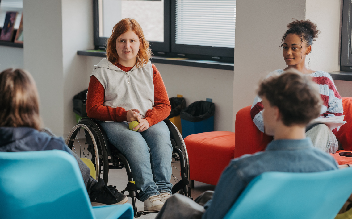 young people sitting in a circle - one white girl in a wheel chair, one black girl with hair up, white person unknown gender with back to view