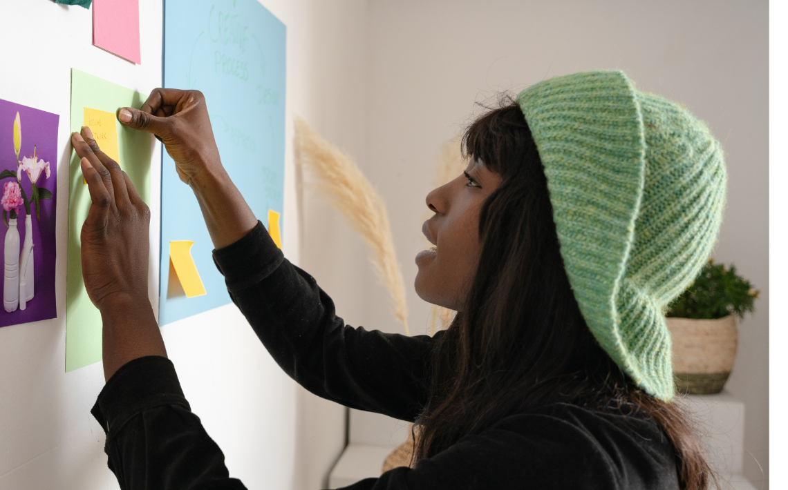 Young person with brown skin and green hat pinning a post it to a wall