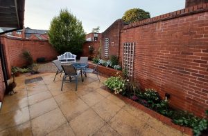 A wide view of the renovated garden at St Luke's hospital, a table and chairs sits in front of a circular space around which flowers have been planted. A large green conifer tree stands in the background. The garden is surrounded by tall red brick walls with trellises.